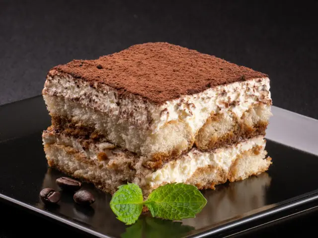 Pyszne tiramisu bez pieczenia, przełożone kremem i biszkoptami, posypane kakao. Obok leżą ziarna kawy i listek mięty.
